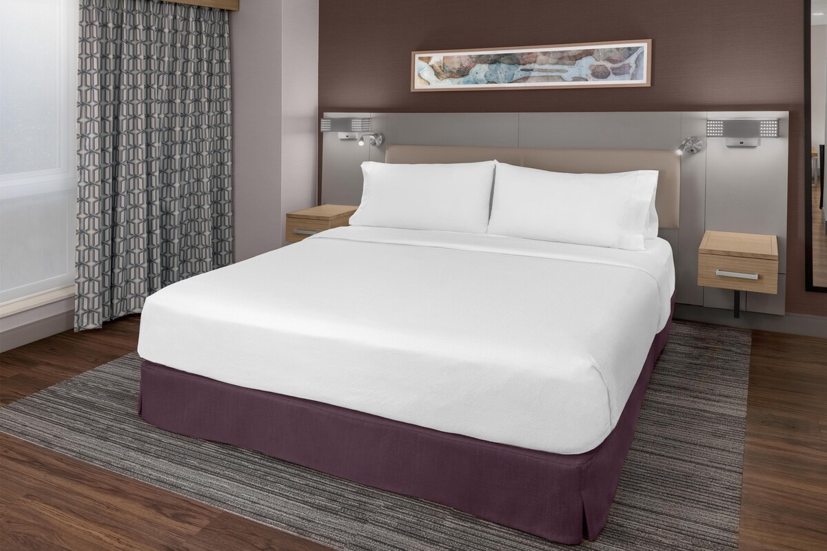 A hotel room with a large bed, a nightstand, a drawer, a headboard with a light fixture, and a framed map on the wall.
