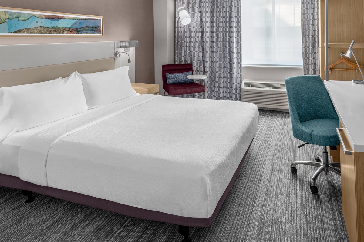 A hotel room with a bed, a desk, a chair, a map on the wall, and a window with curtains.