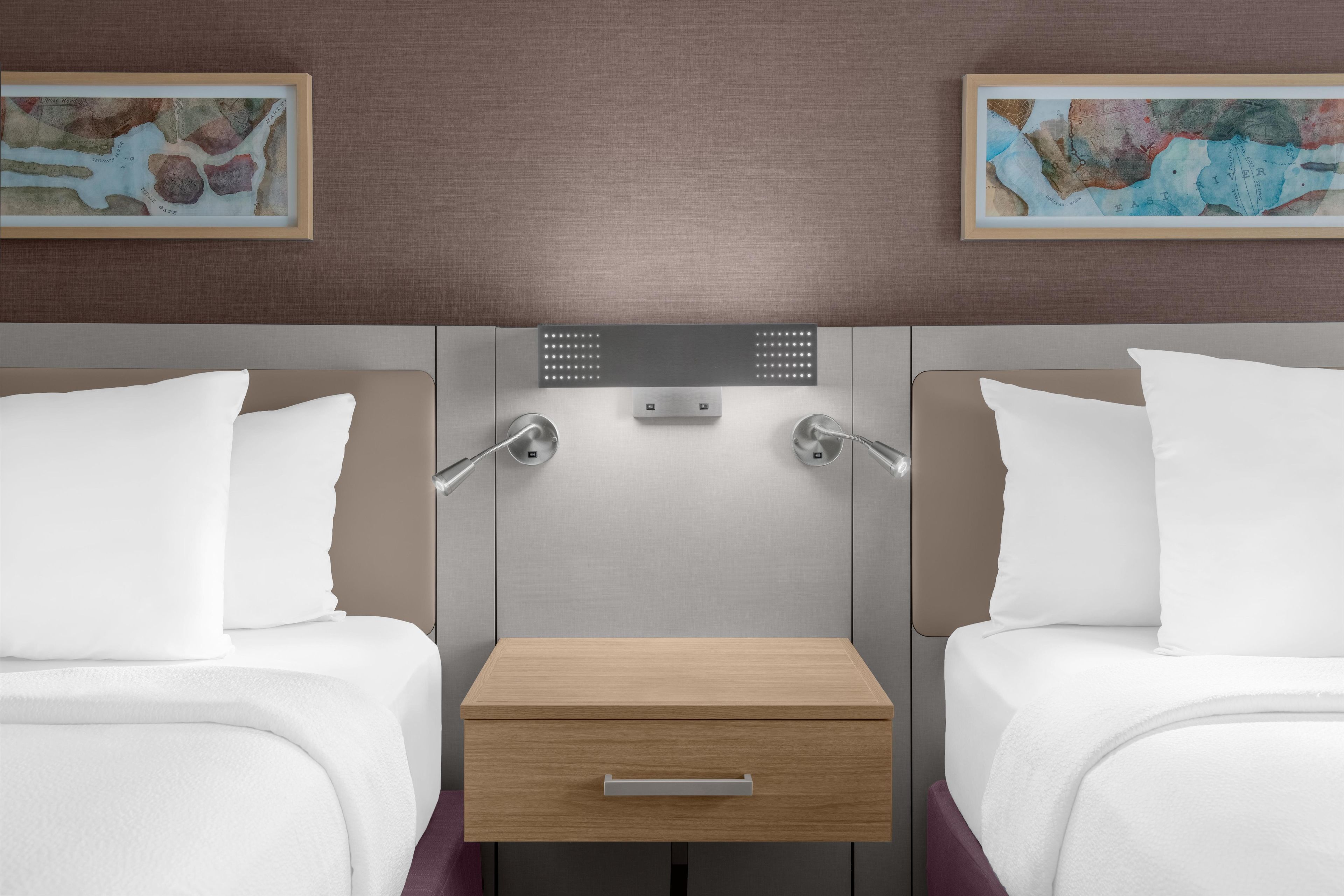Ein Hotelzimmer mit zwei Betten, einem kleinen Nachttisch, einer an der Wand montierten Bedieneinheit und zwei gerahmten Kunstwerken.