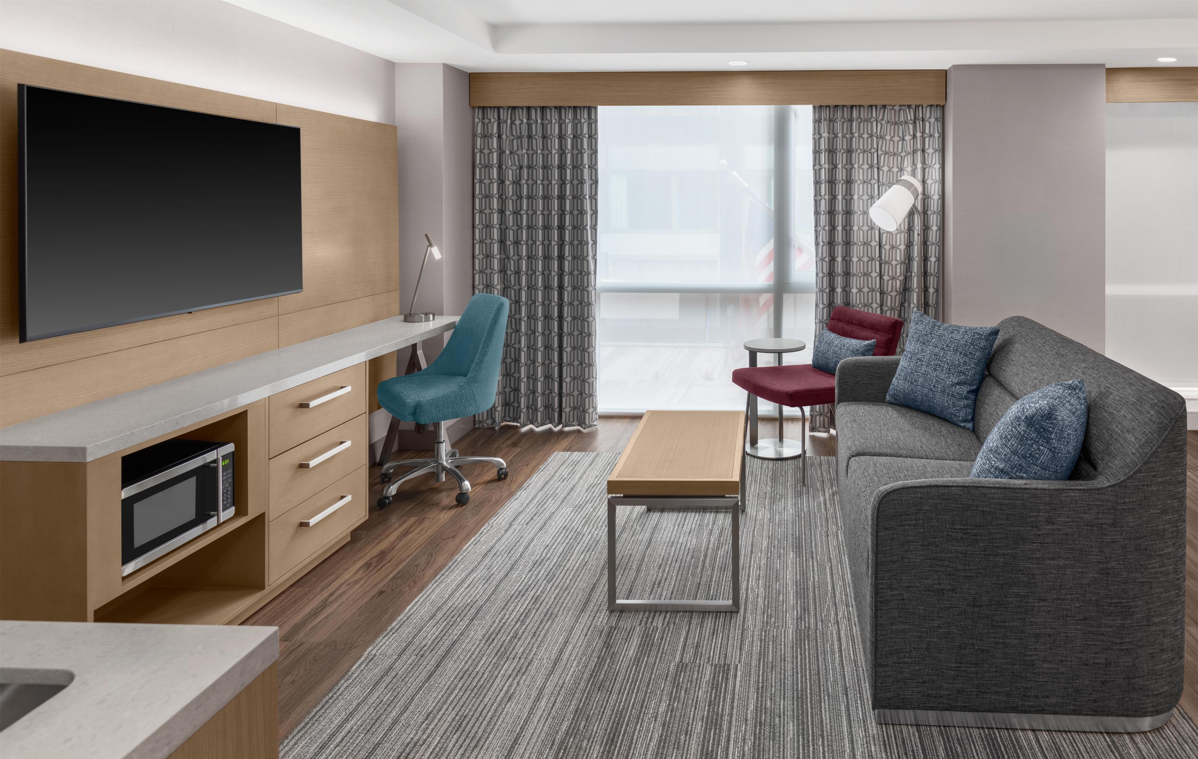 A modern hotel room with a flat-screen TV, a desk with a lamp, a sofa with cushions, and a large window with curtains.