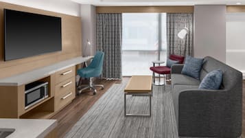 A modern hotel room with a flat-screen TV, a desk with a lamp, a sofa with cushions, and a large window with curtains.