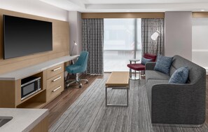 A modern hotel room with a flat-screen TV, a desk with a lamp, a sofa with cushions, and a large window with curtains.