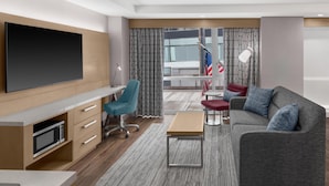 Een moderne hotelkamer met een flatscreen televisie, een bureau met een magnetron, een blauwgroene bureaustoel, een grijze bank met blauwe kussens, een klein houten tafeltje en uitzicht op de stad door grote ramen.