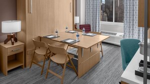Een hotelkamer met een houten bureau, stoelen, een lamp, een raam met gordijnen en uitzicht op de stad.