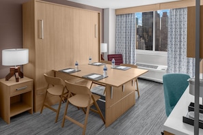 Ein Hotelzimmer mit einem Holztisch, Stühlen, einer Lampe, einem Fenster mit Vorhängen und Blick auf die Stadt.