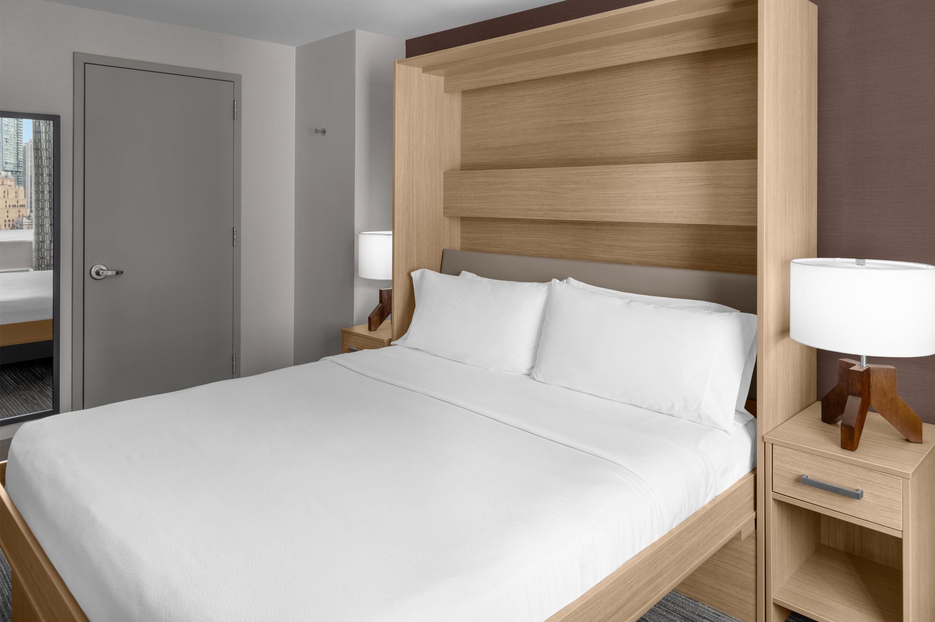Una habitación de hotel con una cama grande, un cabecero de madera, una mesita de noche con una lámpara y una puerta que da a un baño.