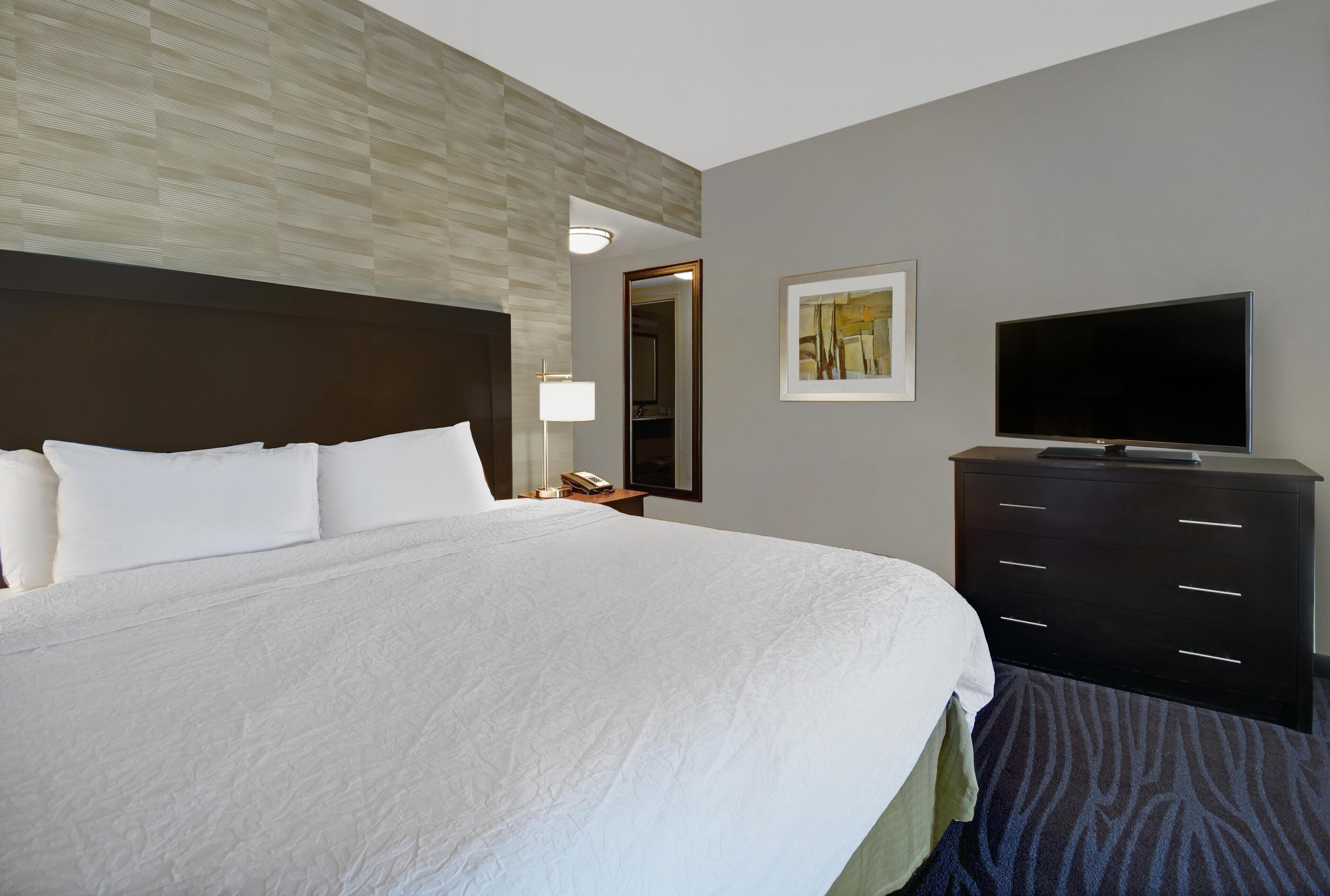 A hotel room with a large bed, a flat-screen TV, a dresser, a nightstand, a lamp, and a framed picture on the wall.