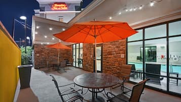 Un área de patio al aire libre con una mesa redonda y sillas, un paraguas grande color naranja, y vista a la piscina a través de amplios ventanales.