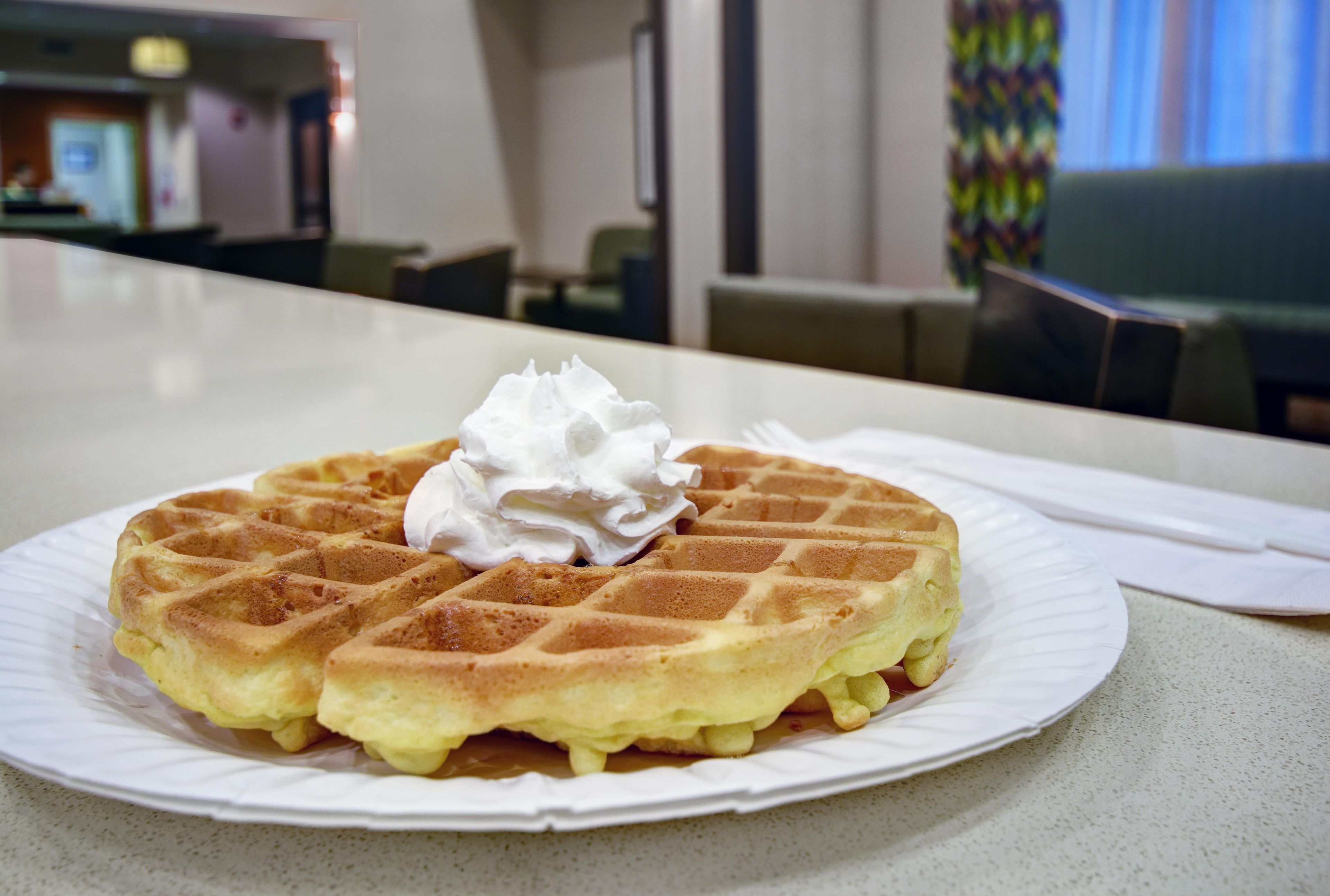 A waffle with whipped cream on a white plate on a table in a room with a view of a cityscape.