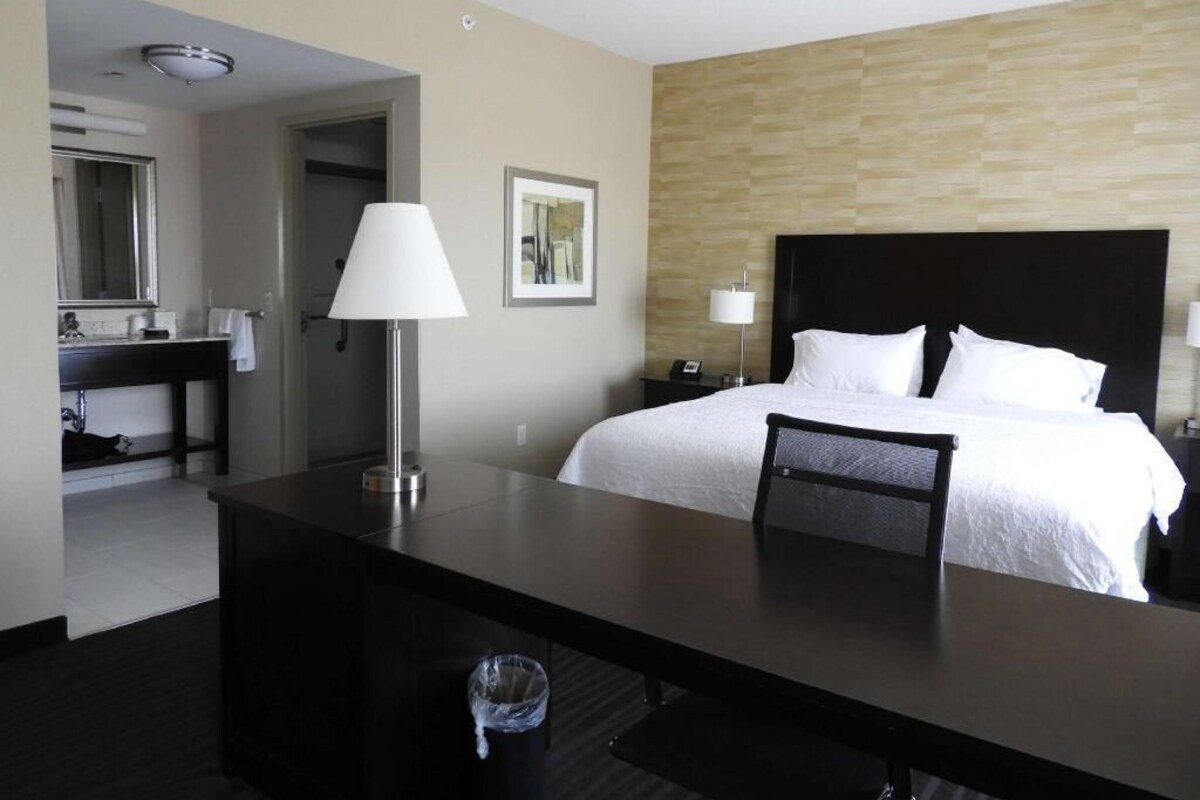 A hotel room with a bed, bedside table, lamp, and a desk with a chair.