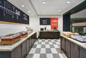 Espaço de buffet de café da manhã com piso xadrez, lousa com desenhos de comidas e placa anunciando waffles.