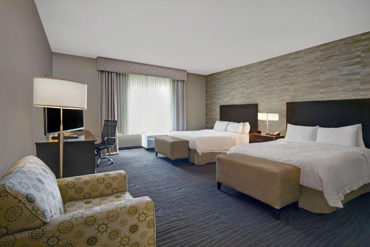 A hotel room with two beds, a desk, a chair, and a large window with curtains.