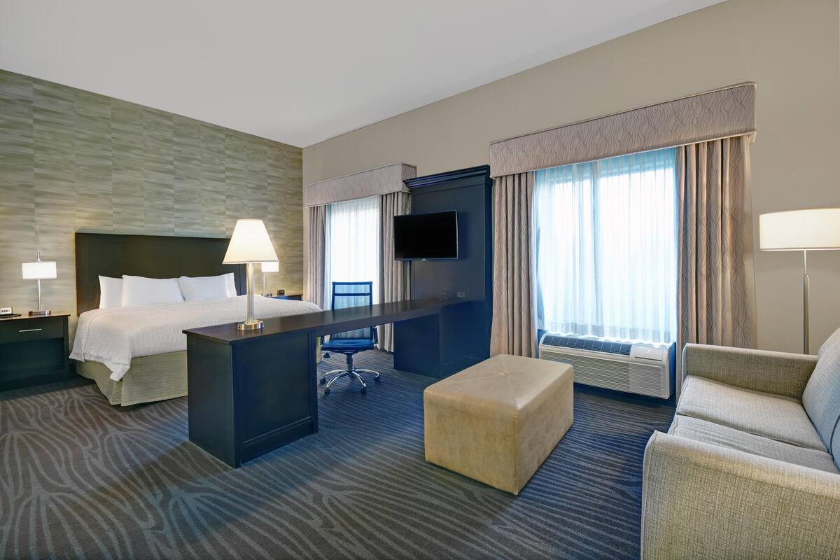 A hotel room with a bed, desk, chair, ottoman, TV, and large windows.