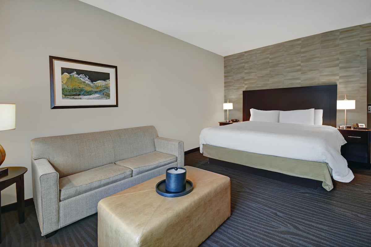 A hotel room with a bed, a sofa, a coffee table, a painting on the wall, and a nightstand with a lamp.