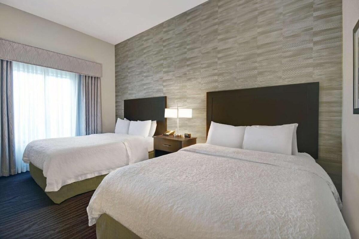 A hotel room with two beds, a nightstand with a lamp, and a window with curtains.