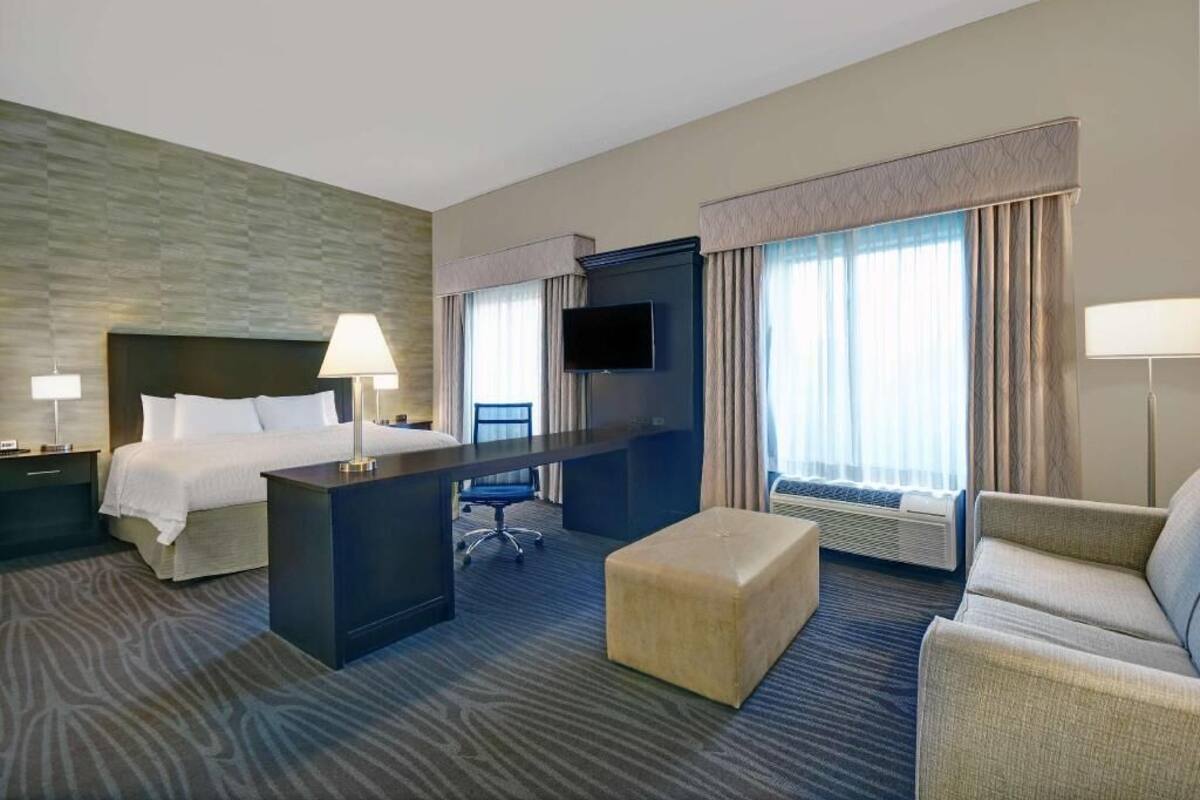 A hotel room with a bed, desk, chair, ottoman, TV, and large windows with curtains.