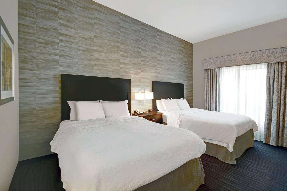 A hotel room with two beds, a patterned wall, a large window with curtains, and a nightstand with a lamp.
