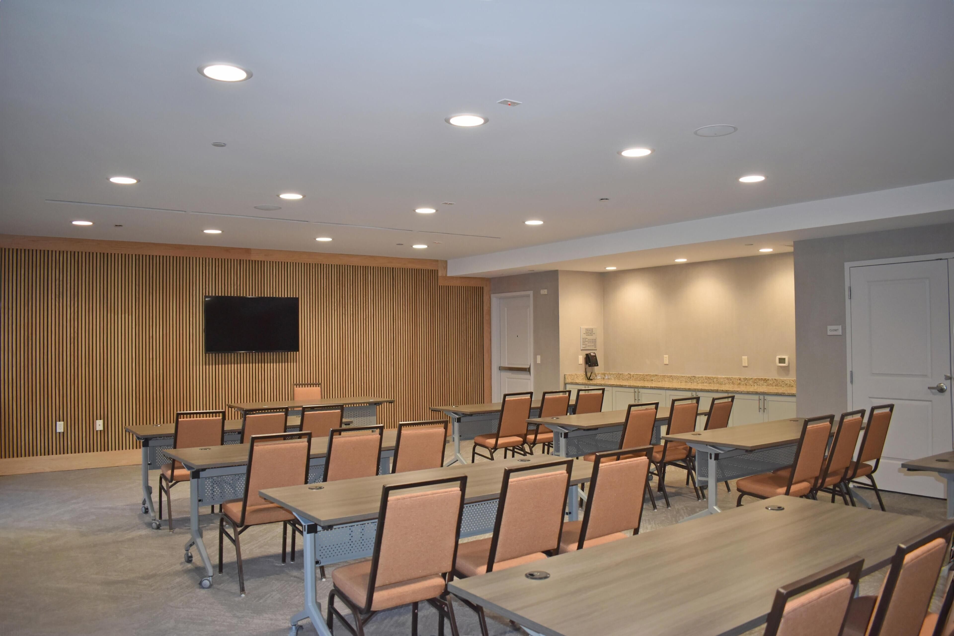 A conference room with tables and chairs, a flat-screen TV, and a kitchenette area.