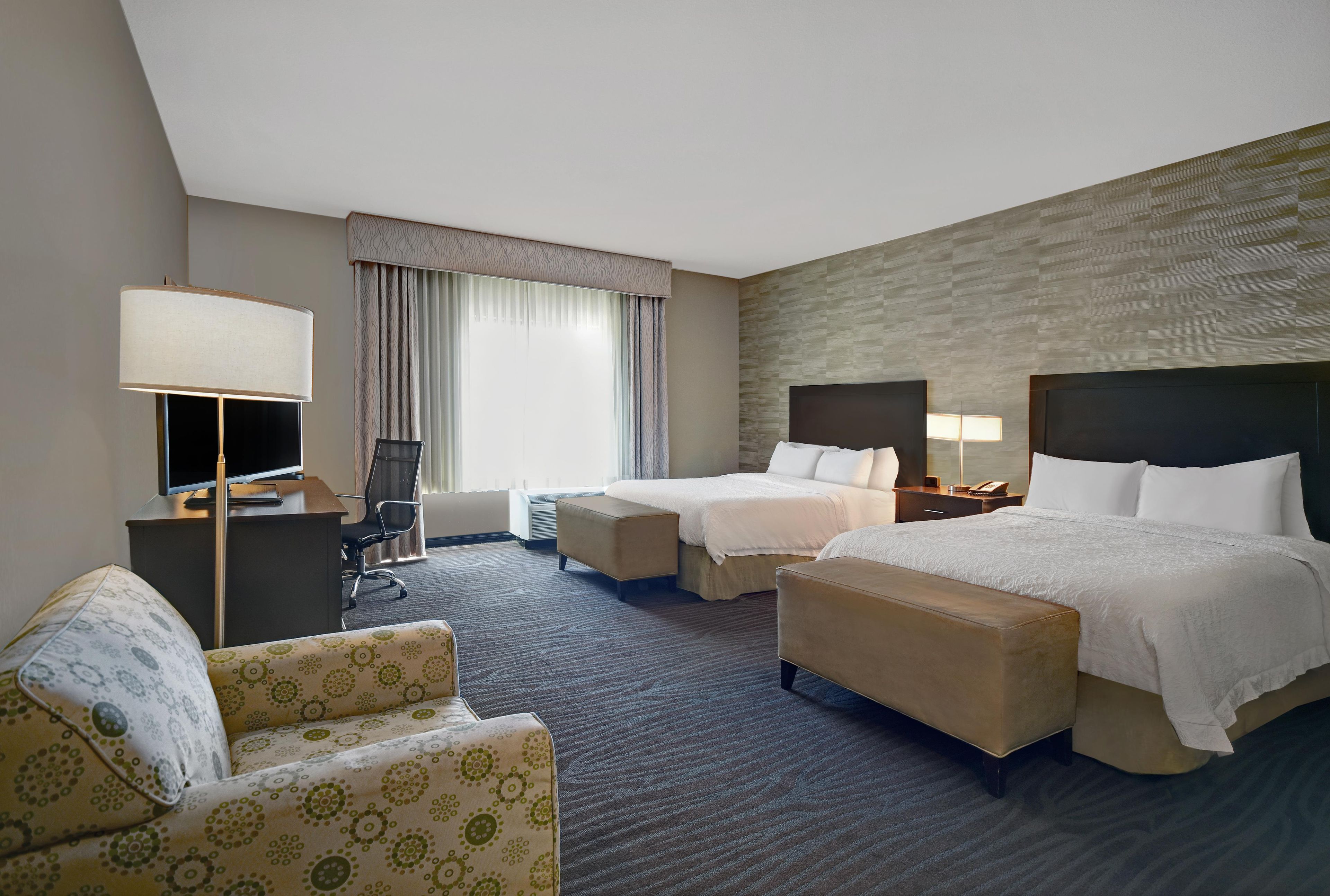 A hotel room with two beds, a desk, a chair, a lamp, and a large window with curtains.