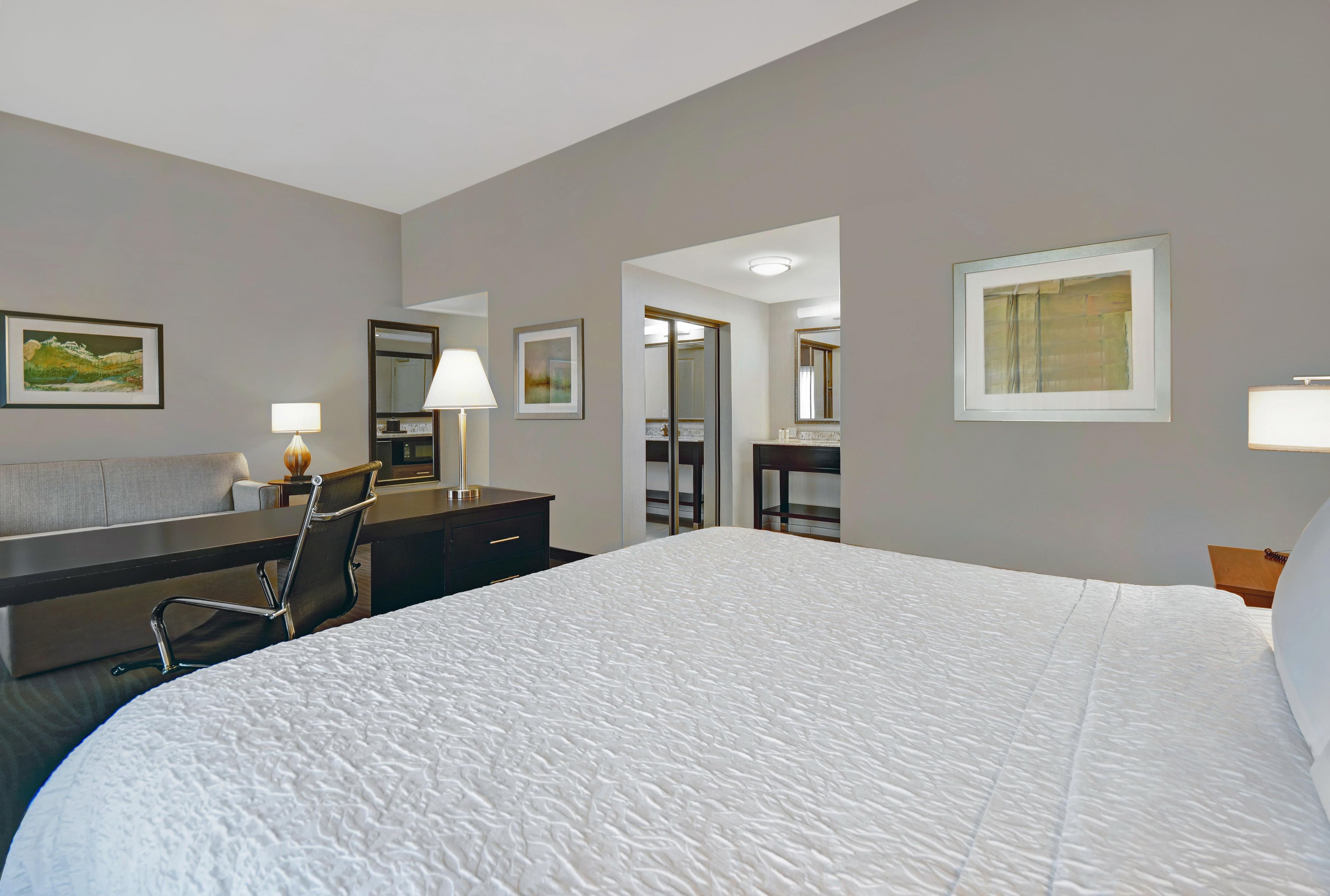 A hotel room with a large bed, a desk with a chair, a mirror, and artwork on the walls.