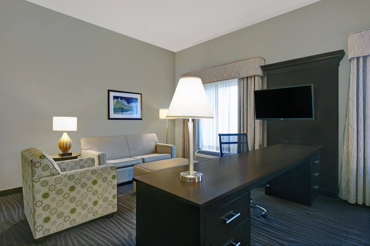 A hotel room with a desk, a sofa, a chair, a TV, and a lamp.