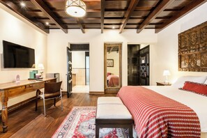 A hotel room with a wooden desk, a chair, a bed with a red and white patterned bedspread, a large mirror, and a decorative wall piece.