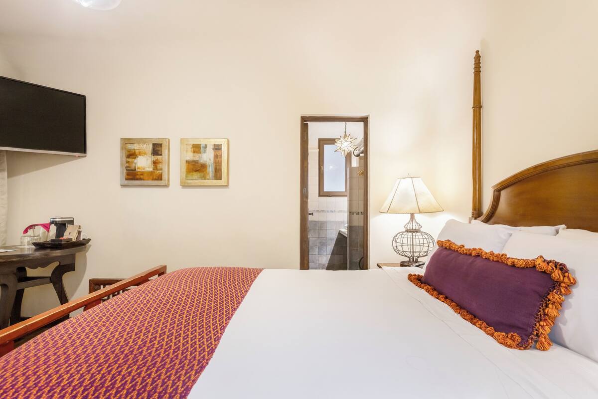 A hotel room with a bed, a desk, a chair, a TV, and two framed artworks on the wall.