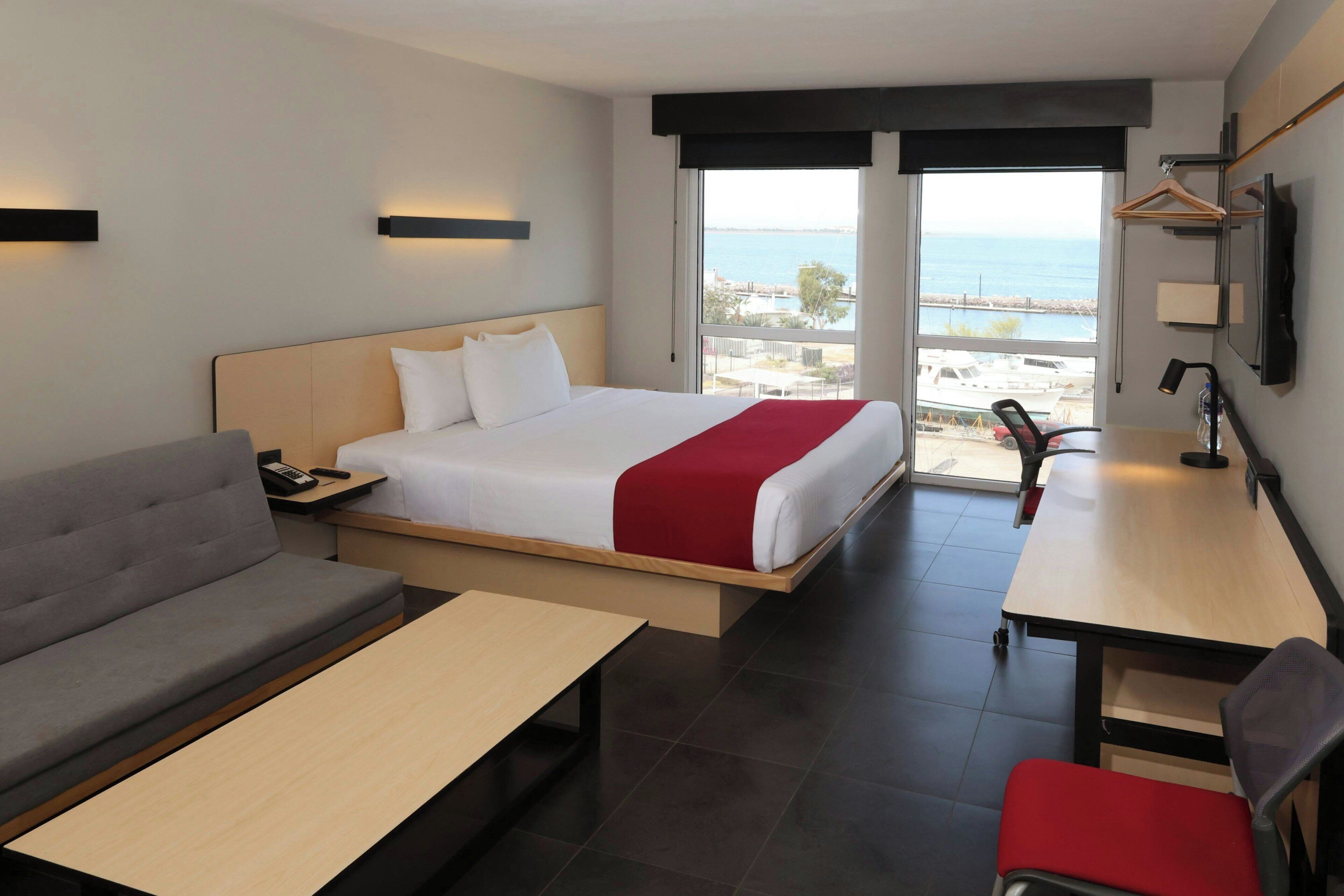 A hotel room with a bed, a sofa, a desk, a chair, a TV, and a large window with a view.