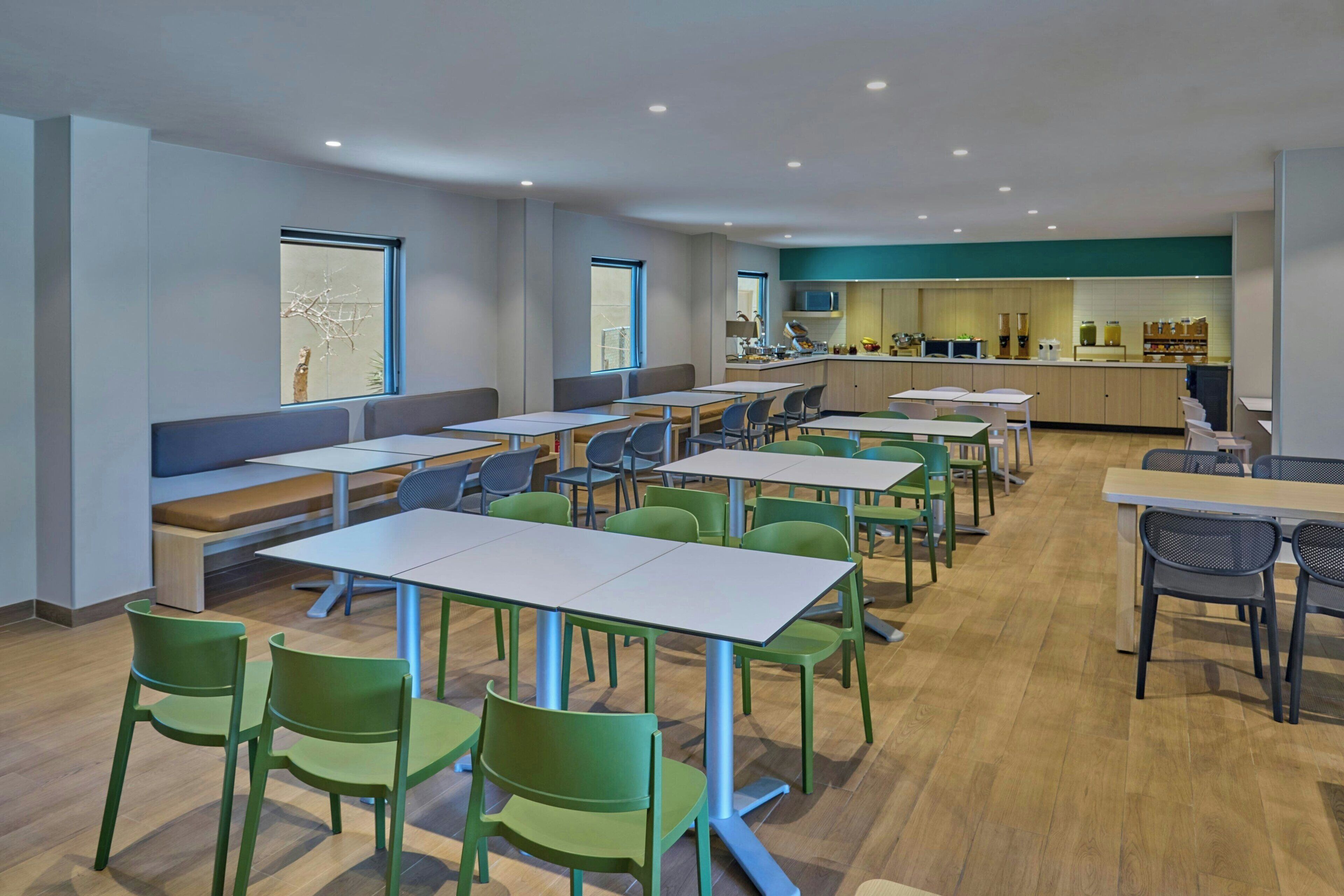 A modern dining area with multiple tables and chairs, a kitchen in the background, and large windows providing natural light.