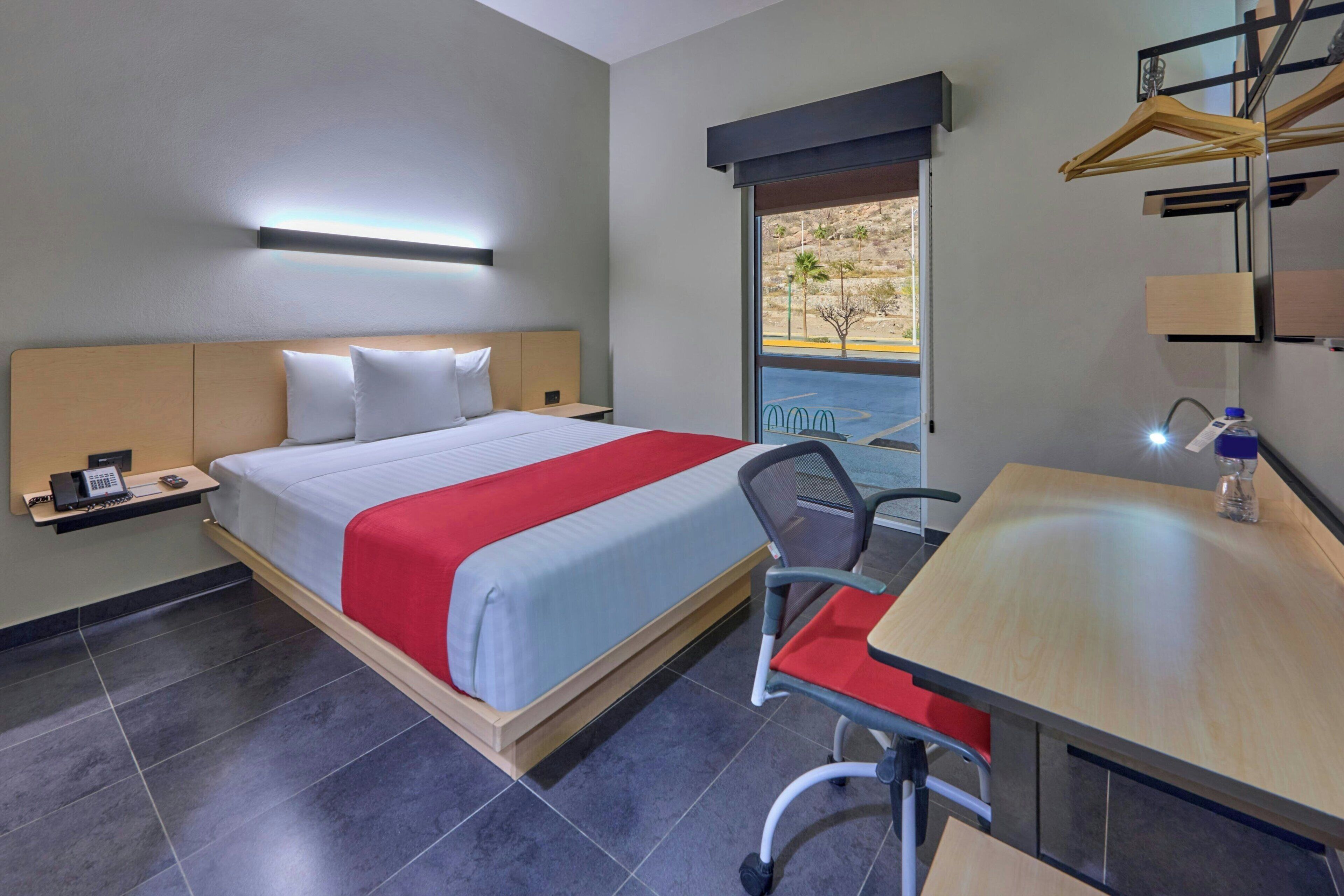 Une chambre d’hôtel équipée d’un lit, d’un bureau, d’une chaise, d’un téléphone et d’une fenêtre donnant sur l’extérieur.
