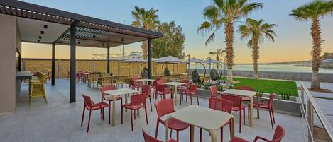 Un patio al aire libre con sillas y mesas rojas y beige, un área de estar cubierta y vistas al mar y palmeras.