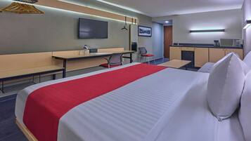 A hotel room with a large bed, a desk with a chair, a kitchenette, and a TV mounted on the wall.