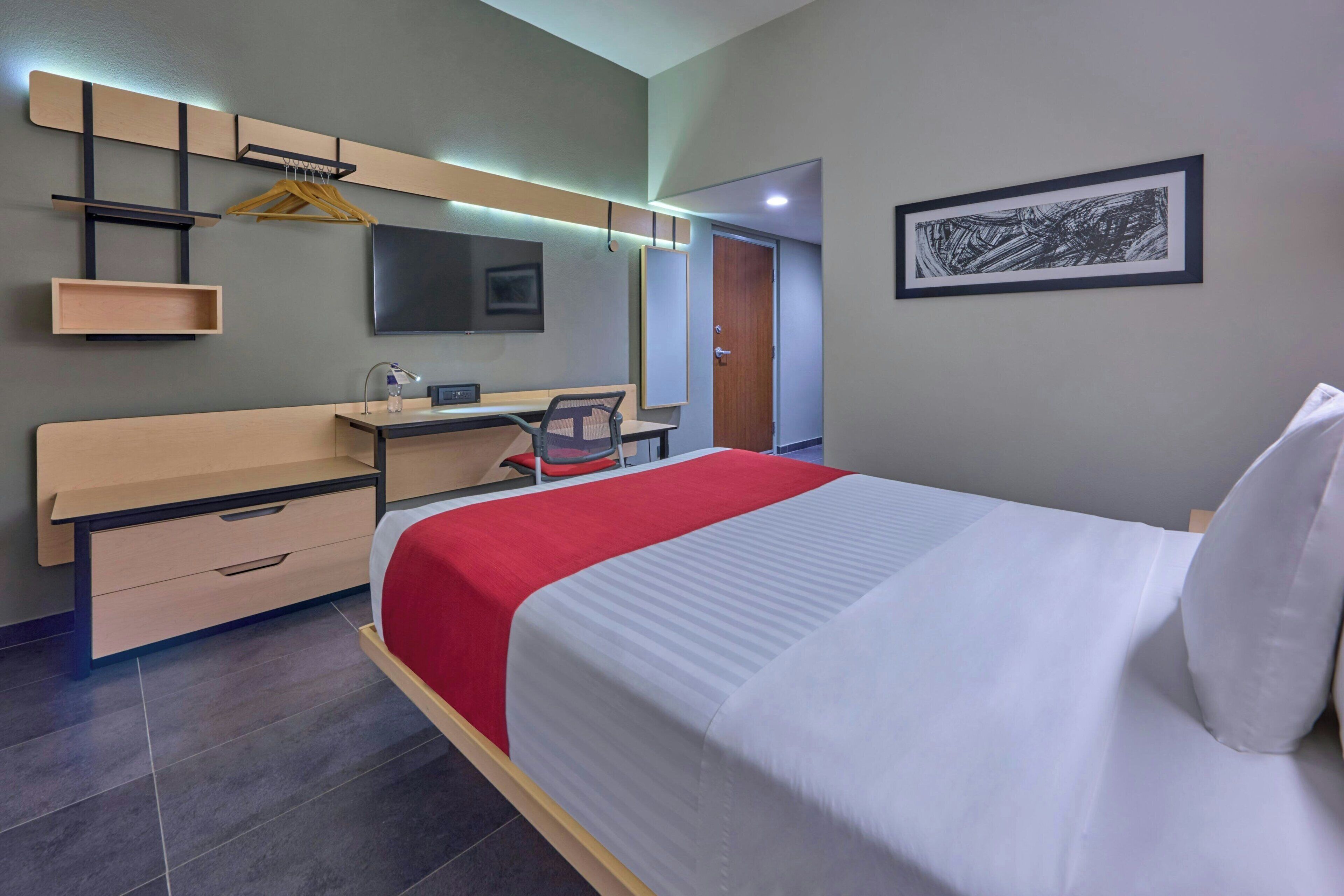 A hotel room with a bed, a desk, a chair, a wardrobe, a mirror, and a framed picture on the wall.