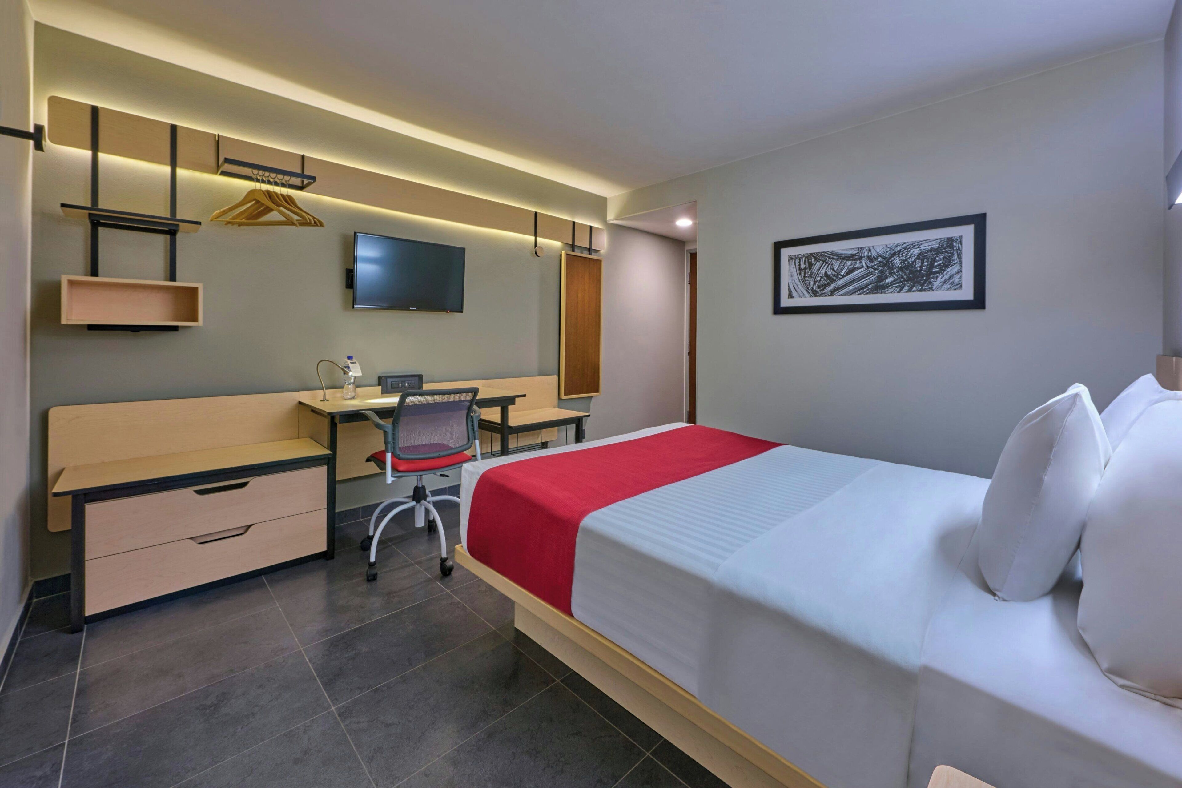 Habitación de hotel con cama, escritorio, silla y televisor de pantalla plana montado en la pared.