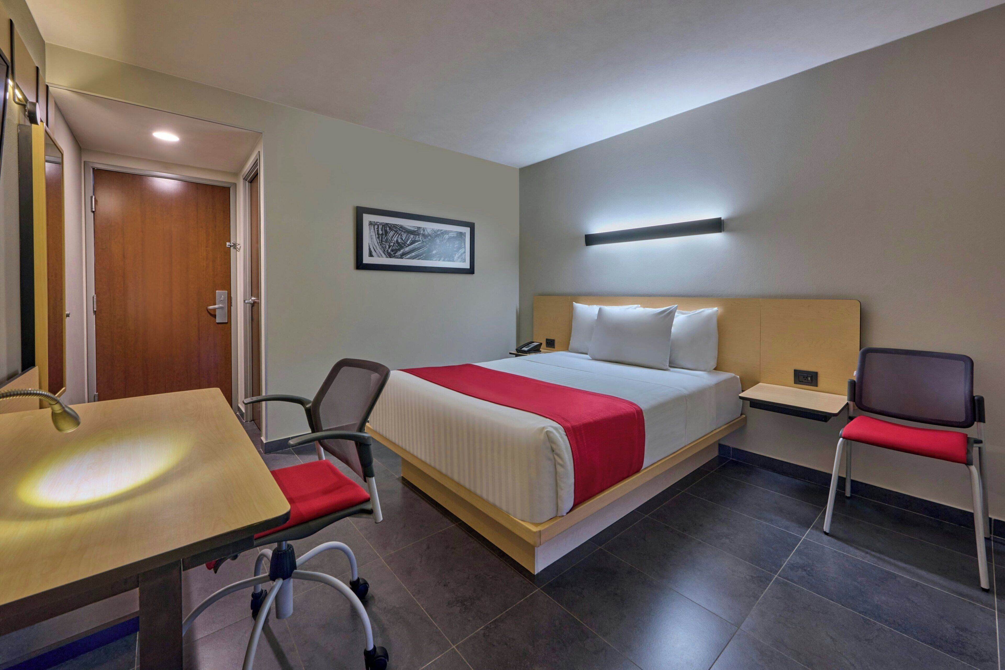 Une chambre d’hôtel comprenant un lit, une table de chevet, une chaise, un bureau avec un lavabo et un tableau encadré au mur.