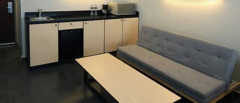 A compact kitchenette with a microwave, sink, and cabinets, a grey sofa, and a small table.