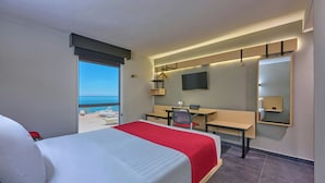 Una camera d'albergo con un letto, una scrivania, una sedia e un'ampia finestra con vista sulla spiaggia.