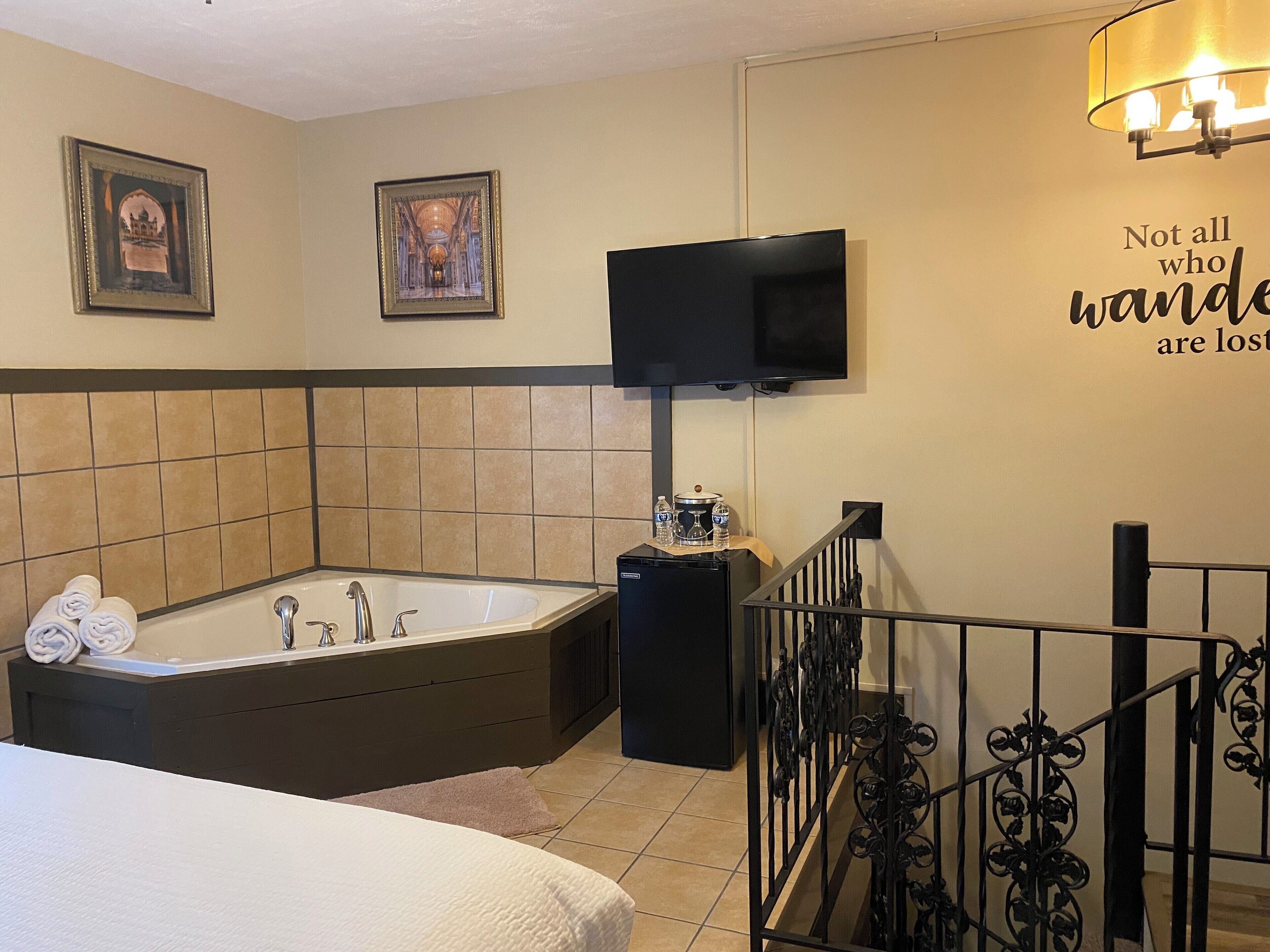 A modern bathroom with a large bathtub, a toilet, and a sink. There are two framed pictures on the wall, a flat-screen TV mounted above the bathtub, and a motivational quote on the wall.