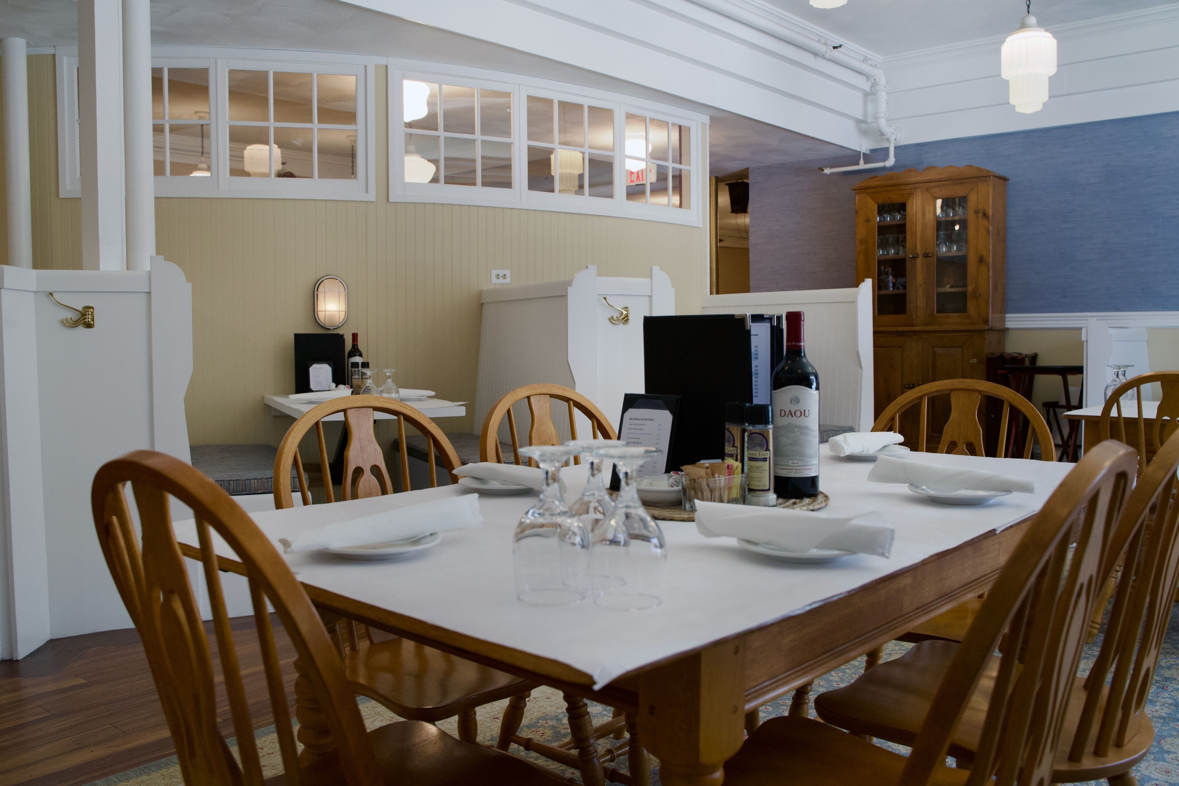 A dining area with a large wooden table set for six, surrounded by wooden chairs. There is a wooden cabinet with glass doors to the right.