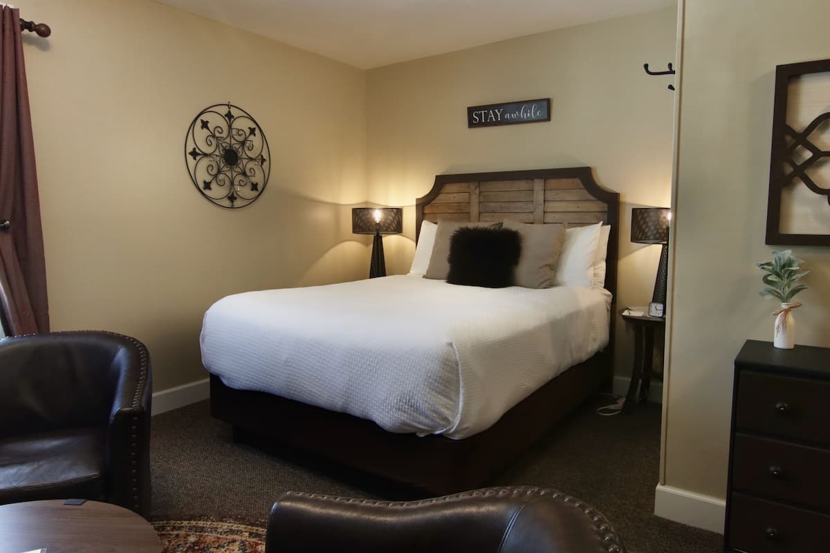 Een hotelkamer met een bed, twee nachtkastjes met lampen, een stoel, een klein tafeltje en een decoratief kunstwerk aan de muur.