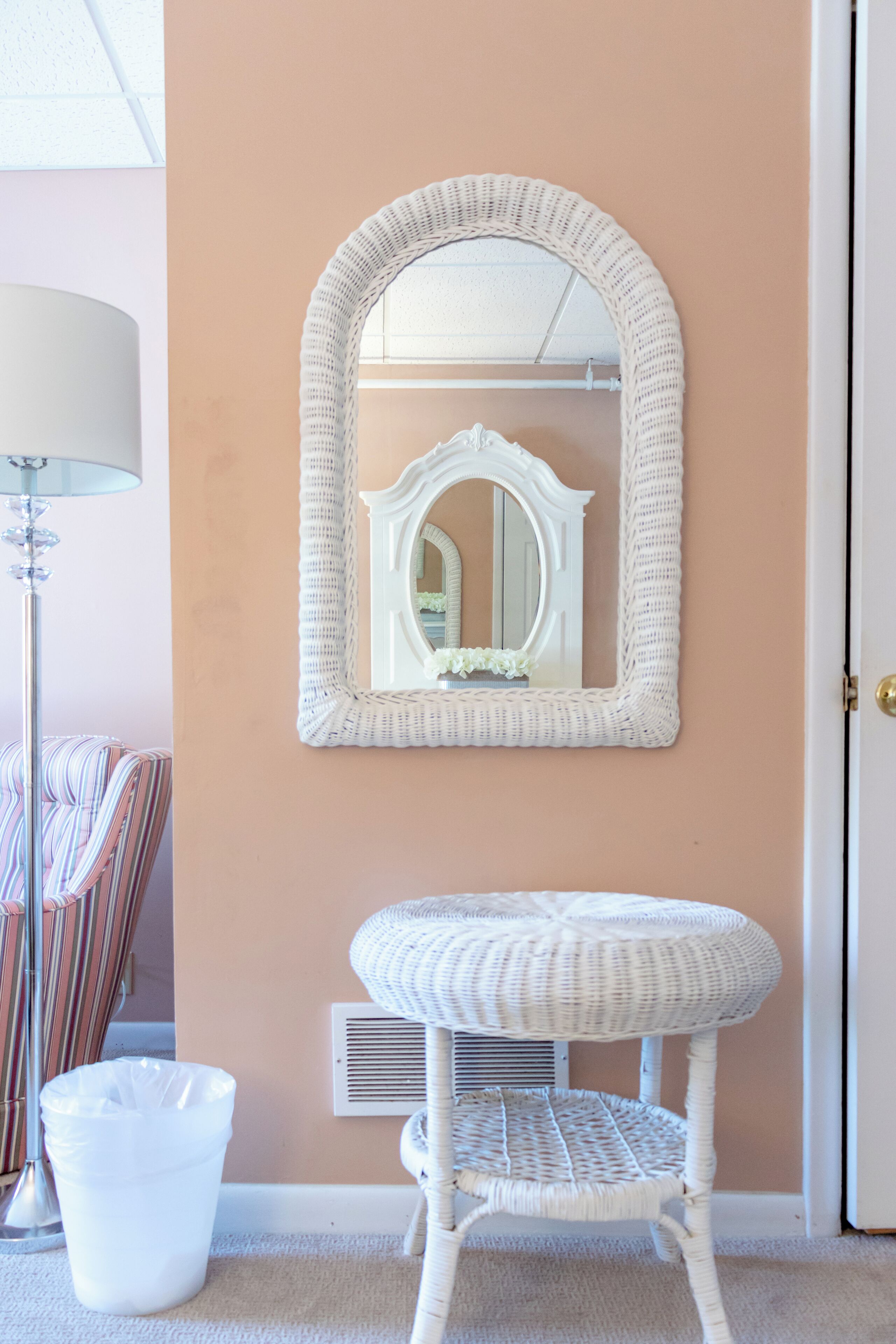 Un baño con un tocador de mimbre blanco, un espejo grande con marco blanco, una silla a rayas y un cubo de basura blanco.