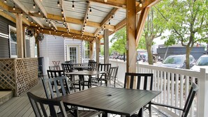 An outdoor patio area with wooden tables and chairs, a covered section with string lights, and a view of the street with cars and buildings in the background.