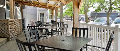 An outdoor patio area with wooden tables and chairs, a covered section with string lights, and a view of the street with cars and buildings in the background.