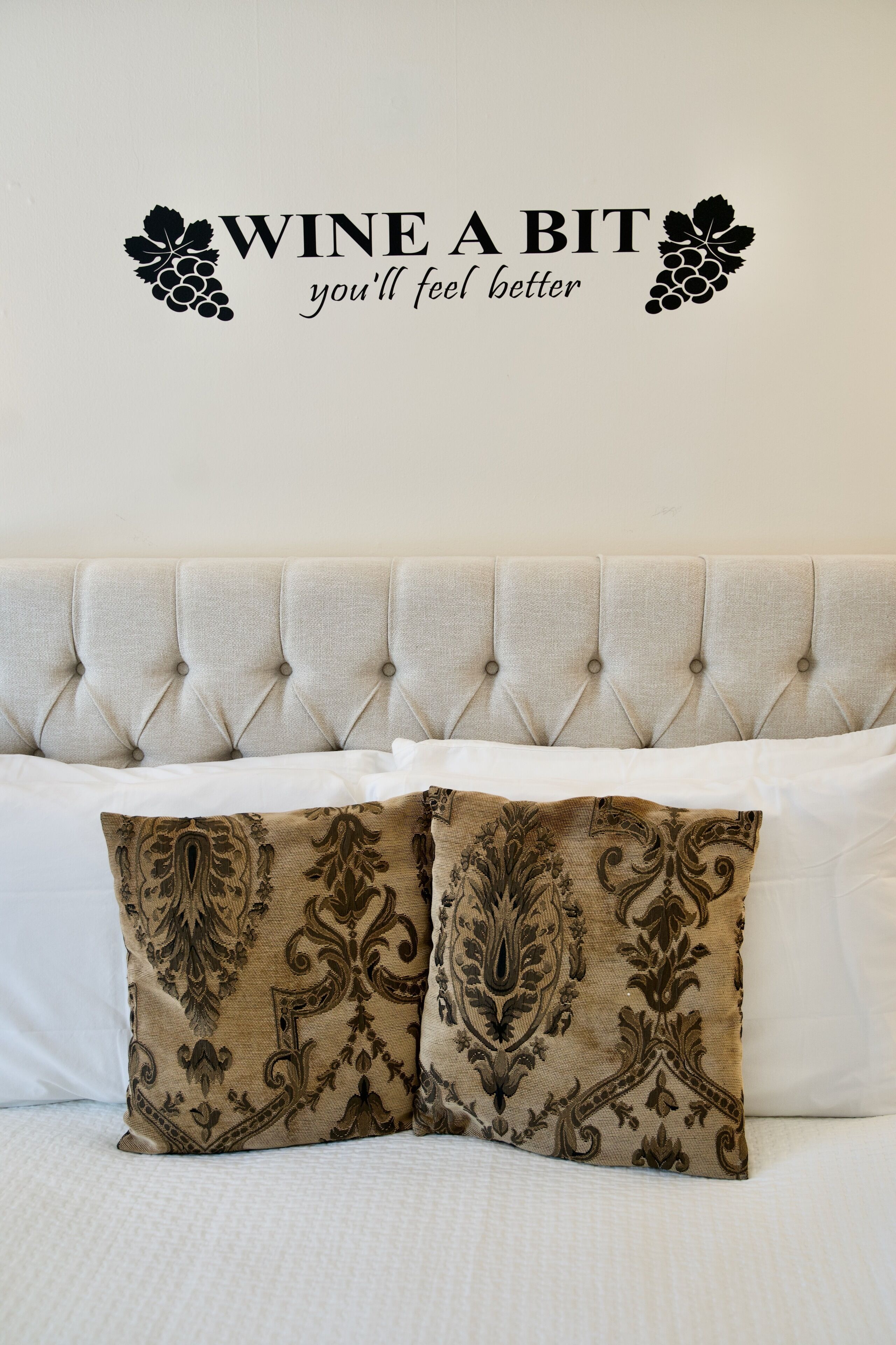 Een bed met een gestoffeerd hoofdbord, twee sierkussens en een muurstick met de tekst 'WINE A BIT you'll feel better'.