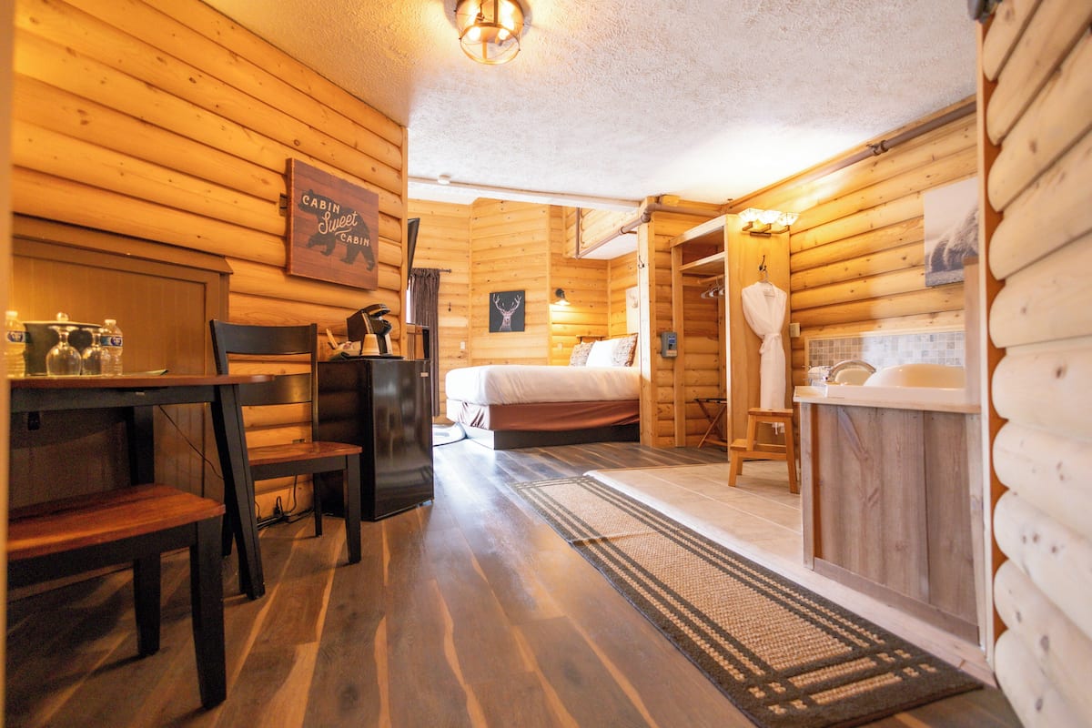 Een blokhutinterieur met een houten vloer, een bed, een tafel met stoelen, een wastafel en een douchehoek.