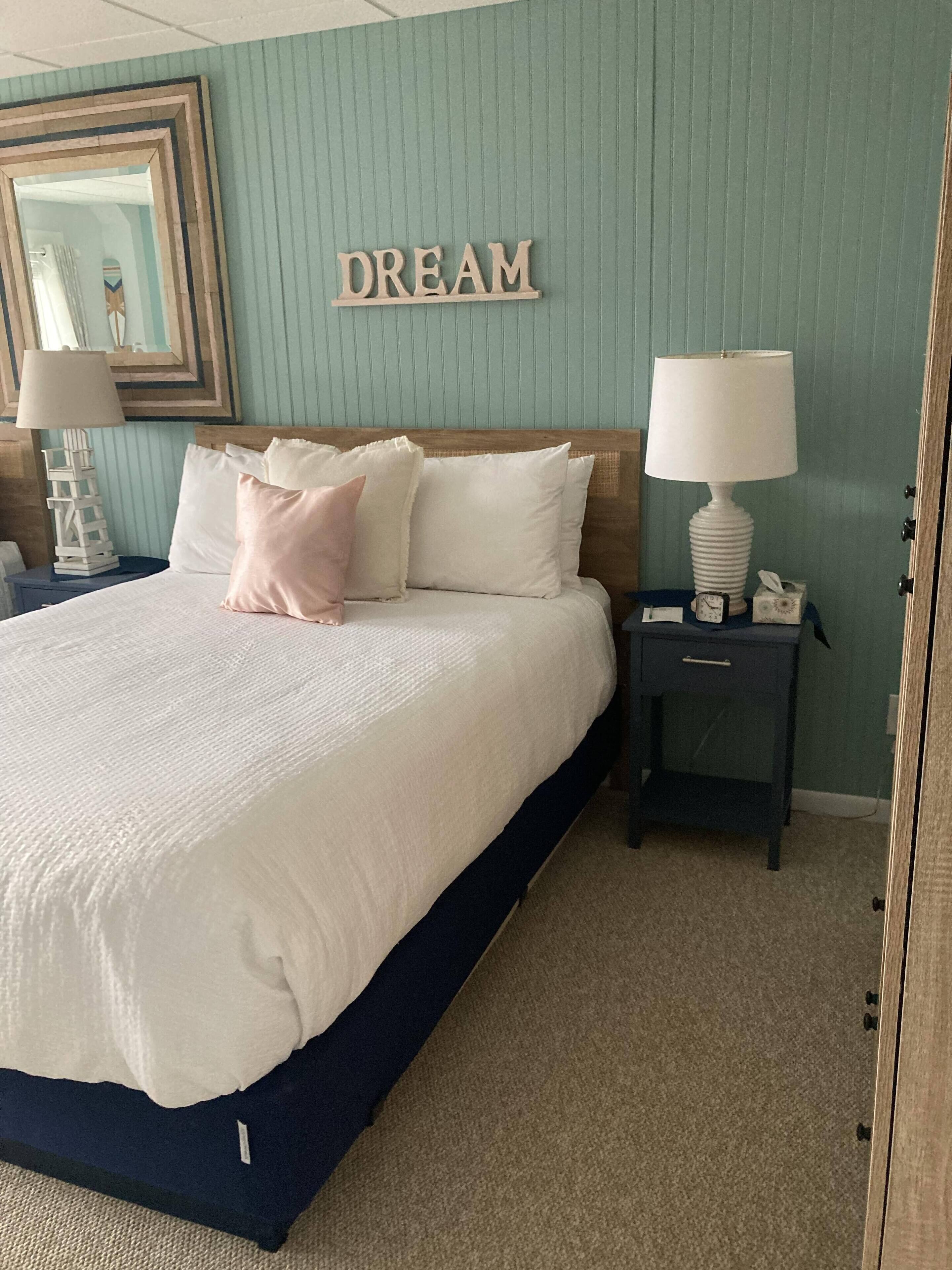 Una cama bien hecha con ropa blanca y almohadas rosas, un cabecero de madera, una mesita de noche con una lámpara, y un cartel en la pared que dice "DREAM".