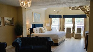 A hotel room with a large bed, two armchairs, a chandelier, and a view of the city through the windows.