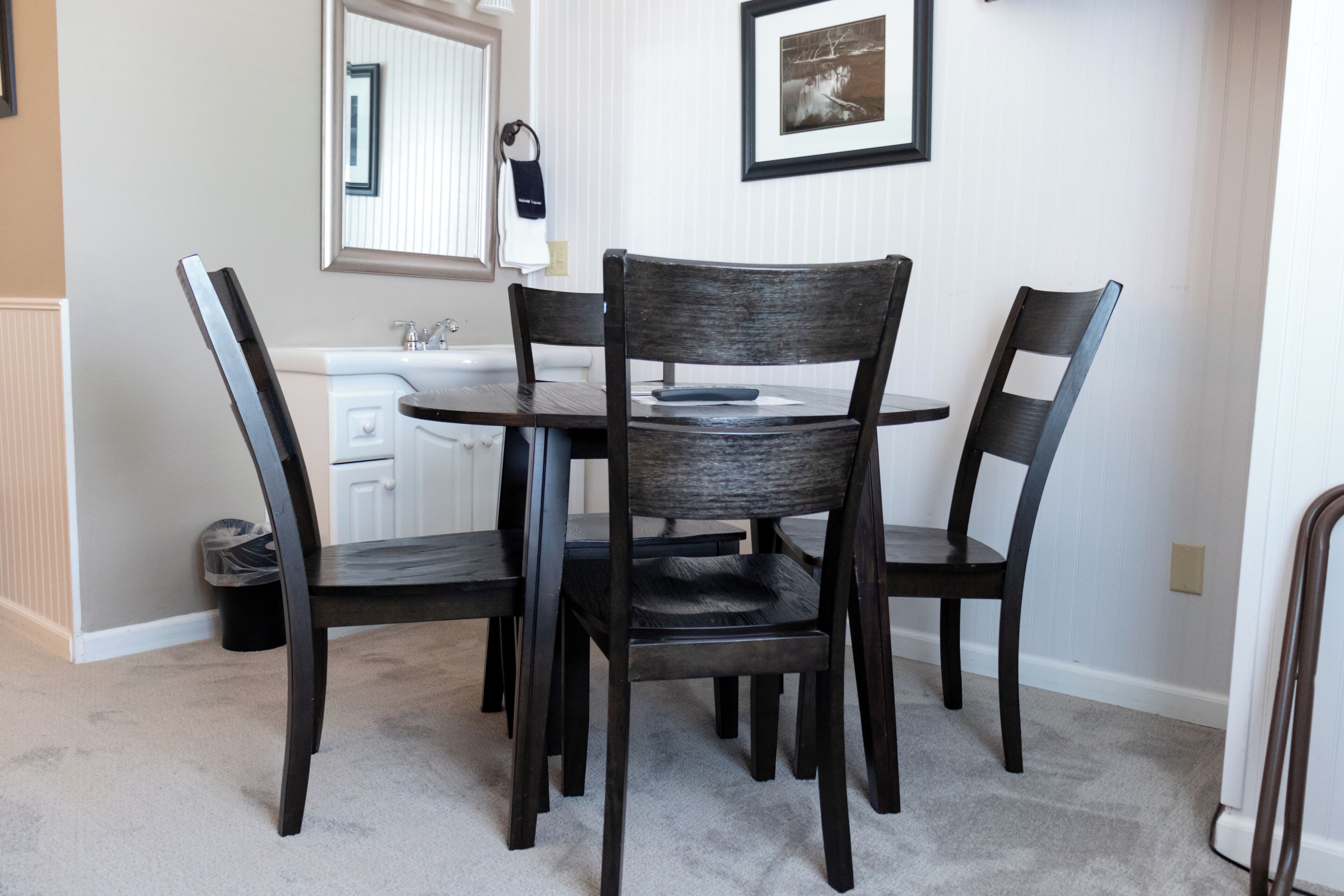 A dining area with a round table and four chairs. There is a white countertop with a sink and a mirror above it. A framed picture hangs on the wall.