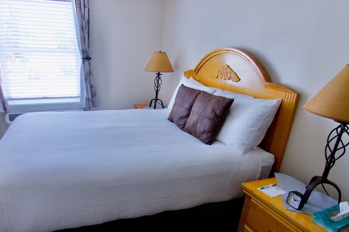 Een netjes opgemaakt bed met een houten hoofdbord, twee bedlampjes, een nachtkastje met een klok en een raam met gordijnen.