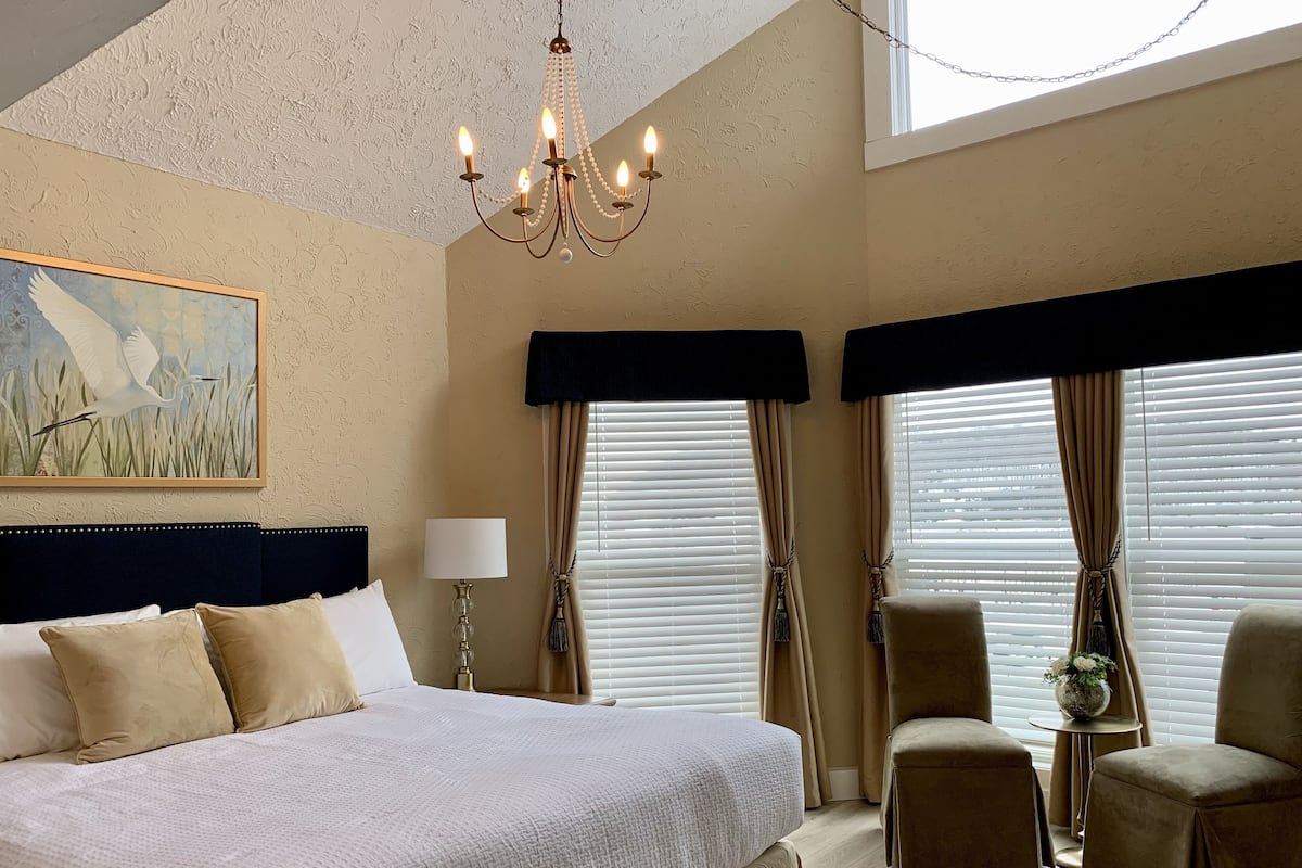Een slaapkamer met een groot bed, een kroonluchter, een schilderij en een zithoek met een stoel en een tafeltje.